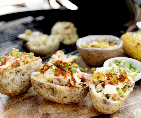 Potato loaded with toppings on a cutting board in front of a grill.